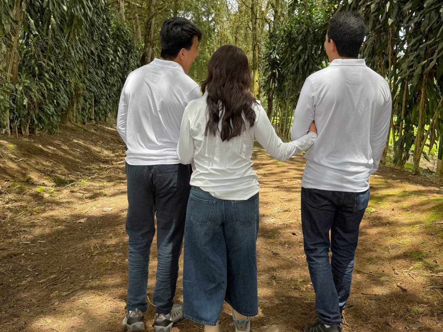 Three people walking away from the camera along a shaded dirt path, arms around each other