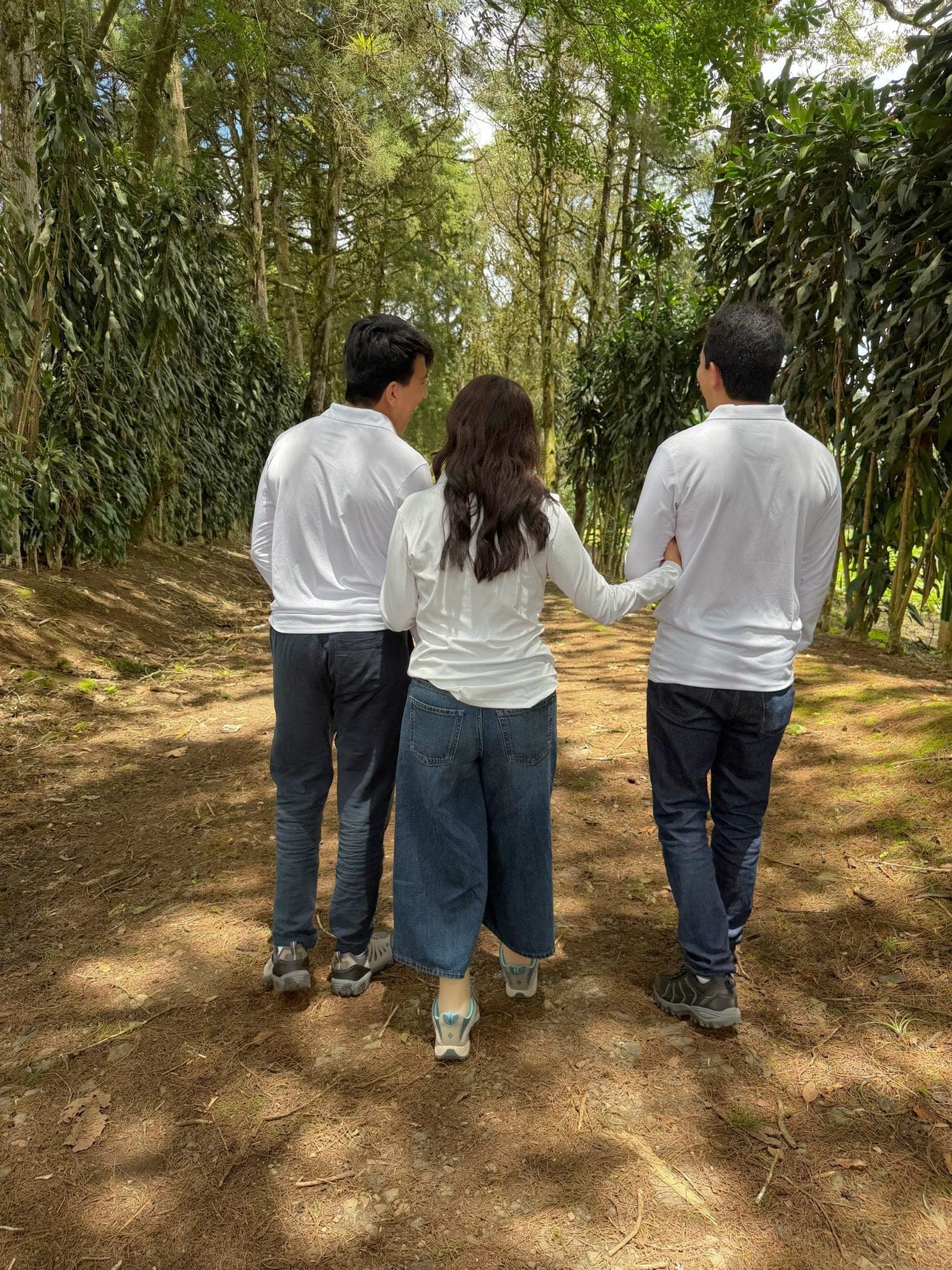 Three adults in white shirts walking away from the camera along a dirt path between trees